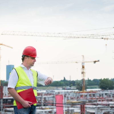 construction worker with a red hard hat checking his watch in front of a large construction site.
